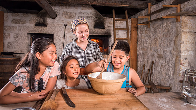 Un enfant mélange de la pâte dans un bol sous le regard et le sourire de deux autres enfants et d’une employée de Parcs Canada en tenue d’époque.
