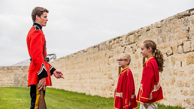 Un employé de Parcs Canada en uniforme rouge de la Police à cheval du Nord-Ouest parle à deux enfants portant le même uniforme.