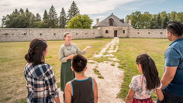 Une employée de Parcs Canada en tenue d’époque s’adresse à quatre visiteurs en faisant un geste vers un bastion de pierre.