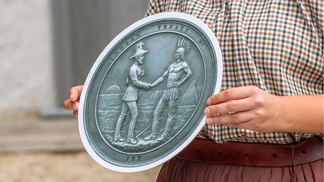 Gros plan d’une employée de Parcs Canada en tenue d’époque tenant une grande plaque métallique représentant un colon et un autochtone se serrant la main, avec le soleil levant et des tipis en arrière-plan.