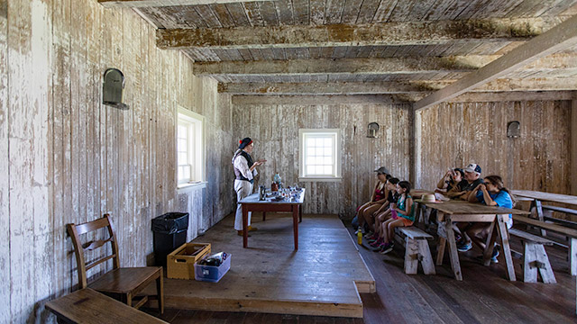Un employé de Parcs Canada en tenue d’époque se tient derrière une table sur laquelle sont posés des artefacts et il s’adresse à un groupe de visiteurs assis sur des bancs.