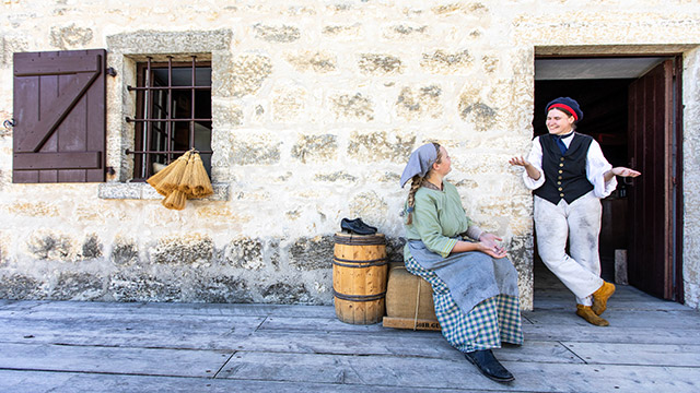 Deux employées de Parcs Canada en tenue d’époque discutent. L’une d’elles est adossée à une porte en pierre, tandis qu’une autre est assise sur une balle de fourrure.
