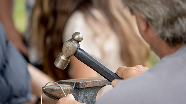 Gros plan d’un visiteur utilisant un marteau pour plier une fine pièce de métal à l’aide d’une enclume.