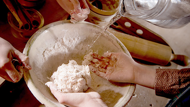 Gros plan d’un bol avec une paire de mains tenant une boule de pâte, une autre paire de mains présentant des morceaux de pâte collés, et de l’eau coulant d’un pichet.