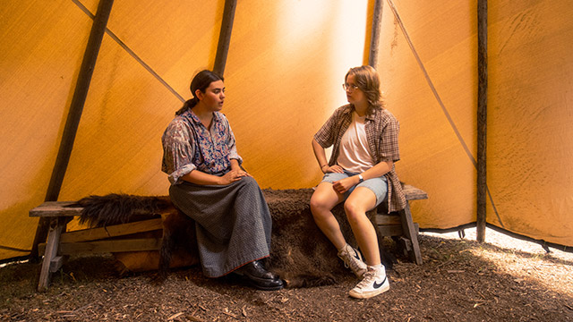 Une employée de Parcs Canada en tenue d’époque parle à une visiteuse alors que toutes deux sont assises sur un banc à l’intérieur d’un tipi.
