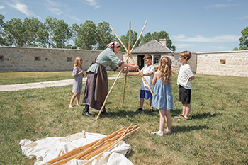 A woman in a historic costume helps a group of children assemble a tipi at Lower Fort Garry.