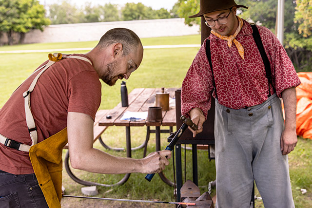 A person hammers a hot metal bar against an anvil as a Parks Canada employee in historic clothing watches them.