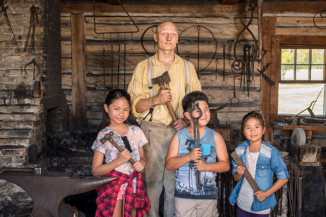 Trois enfants visitant Lower Fort Garry posent pour une photo avec des outils de forgeron et un employé de Parcs Canada en tenue historique.