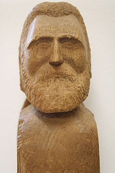 Front view of wood carving of Louis Riel, with the name “David” visible at the base of the bust. The bearded figure rests on a three-piece supporting shelf.