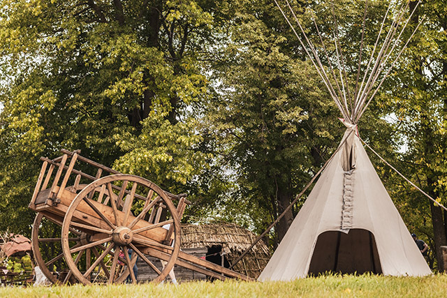 A cart and tipi stand next to some trees with a wigwam in the background