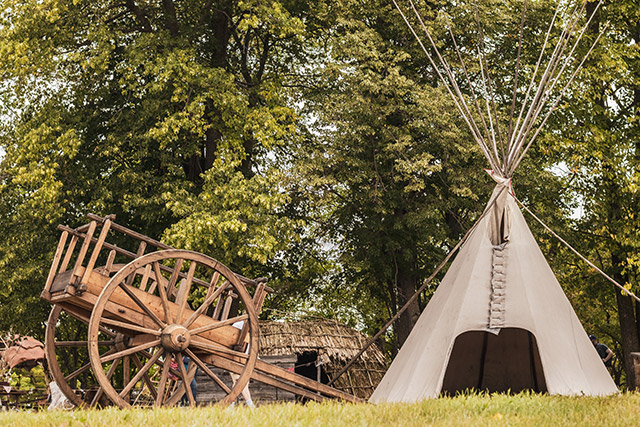 Une charrette et un tipi se trouvent à côté de quelques arbres, avec un wigwam en arrière-plan.