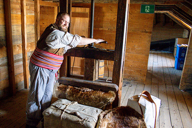 A Parks Canada employee in historic clothing uses a machine with a crank to press down fur pelts.