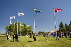 Nine flag poles in a C shape are shown with a flag raised to the top. A person is at the base of each flagpole and a crowd of people is watching at the right of the photo.