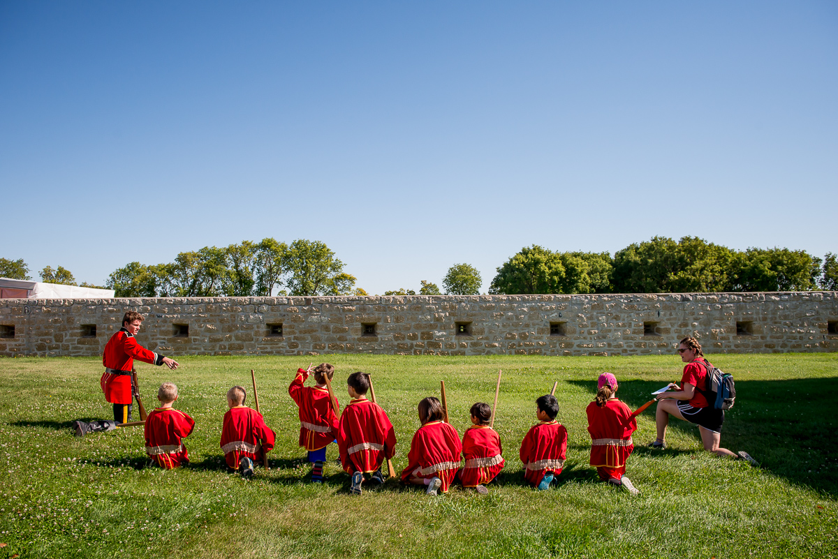 Nine children wearing red uniforms are shown kneeling in the grass looking at a Parks Canada employee, also wearing a red uniform.