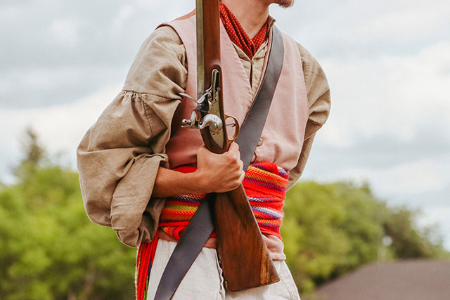 Gros plan du torse d’un employé de Parcs Canada portant des vêtements historiques, dont une ceinture fléchée, et tenant un fusil pointé vers le haut à Lower Fort Garry.