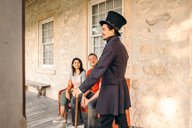 A Parks Canada employee in historic clothing, including a beaver pelt hat, smiles and looks off the Big House porch as two visitors look at him at Lower Fort Garry.