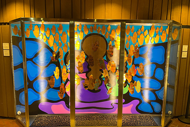 An art piece on display with a painting of a woman and two children, and many paper handprints hanging from the top.