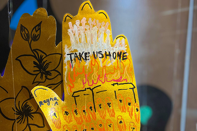 A gold, paper hand with the words “Take us Home’ along with a drawing of orange ghost-like people.