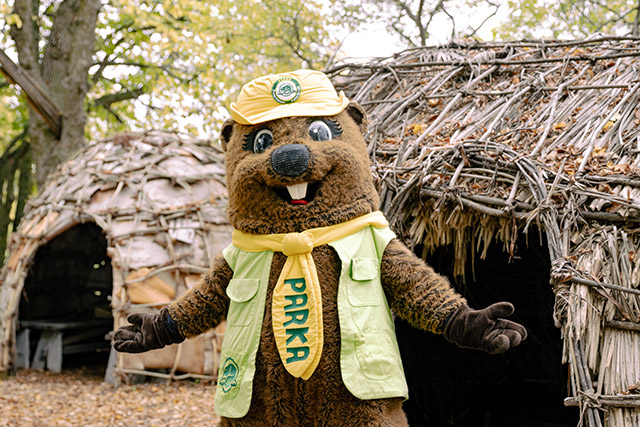Parks Canada beaver mascot Parka gestures to two wigwams in the background.