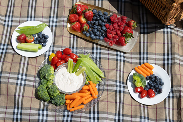 Plates of fruit, vegetables and dips on a picnic blanket.