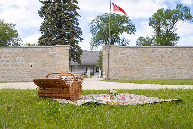 A picnic basket with teacups and platters of sandwiches and desserts on a blanket on the lawn in front of the walls at Lower Fort Garry.