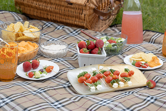 Bowls of chips, dip and berries, platters of fruits and vegetables and glasses of juice on a picnic blanket.