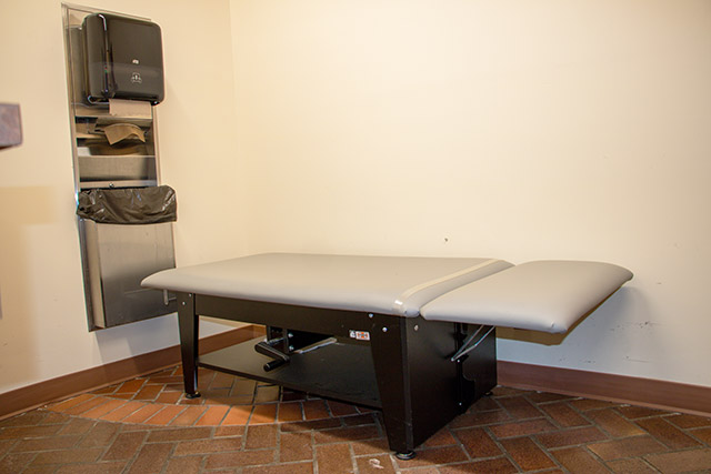 A grey padded table with a headrest. A paper towel dispenser and garbage receptacle are also visible.