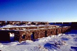 A stone wall at the back of the image serves as an exterior wall for Prince of Wales Fort, while two shorter stone walls closer to the viewer are interior walls with doorways and windows. There is snow on the ground.