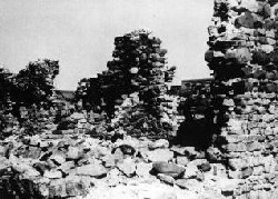 A black-and-white image shows stones in the foreground. At right, there is a brick wall with a jagged edge, and there is a section of brick wall in the middle of the photo further from the viewer.