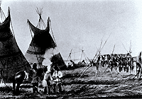 Image en noir et blanc d’un groupe de personnes debout et d’un cheval, au premier plan; à l’arrière-plan, deux tipis et un support fait de bâtons de bois sur lequel des fourrures sont accrochées 