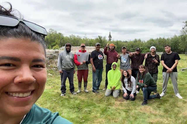 A person takes a selfie as a group of 10 people stands and kneels in the grass in the background.