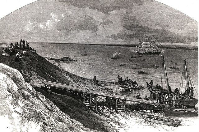 A sloping riverbank with a dock leading down to two sailboats. A larger ship is seen on the water.
