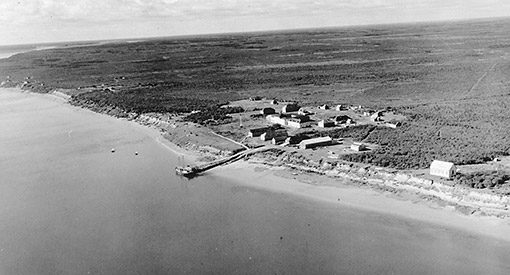 An aerial view of roughly a dozen buildings with a dock leading to the water at left.