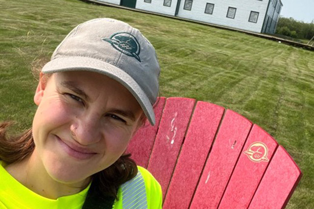 A Parks Canada employee takes a selfie while sitting in a Red Chair with the large, white York Factory depot building in the background.