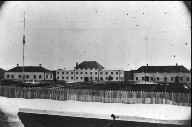 A black-and-white image of three buildings, with a tall fence in the foreground.
