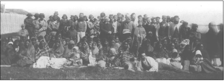A black-and-white image of a large group of Caucasian and Indigenous people posing for a photo. In the left of the image, there is a fence and a rooftop visible.