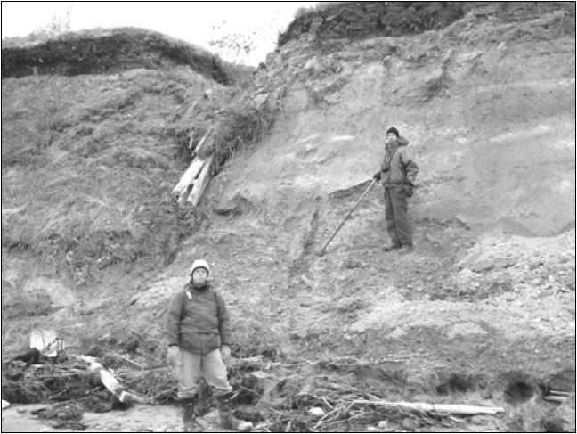 Une personne se tient debout au pied d’une berge abrupte et une autre se tient debout sur la pente de la berge tenant un long bâton.