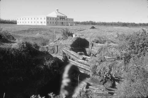 In the foreground, wooden boards are embedded in a trench in the ground. In the background, there is a large white building.