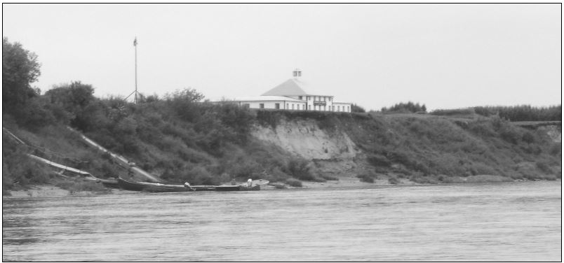 Un grand bâtiment blanc occupe le centre de l’image. Une berge abrupte mène à la rivière. Un quai se trouve à gauche de l’image.