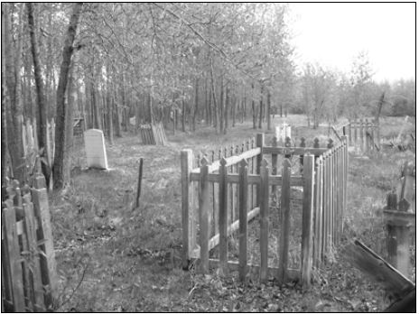 Une pierre tombale entourée d’une clôture en bois. Des arbres bordent le cimetière à gauche de l’image.