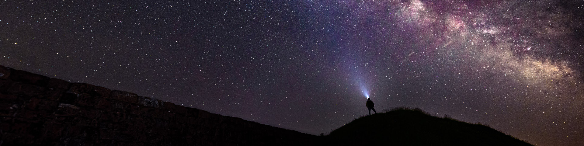 A person wearing a headlight looks at the milkyway on top of a hill at the Fort.