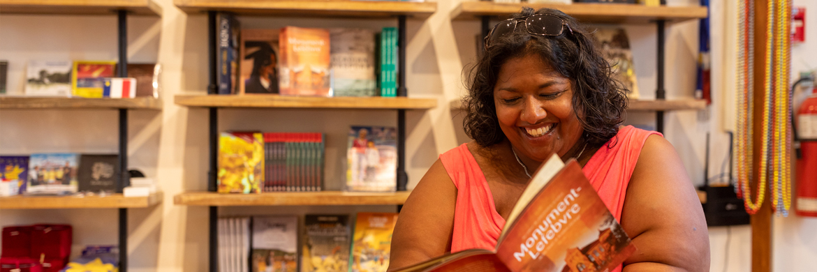 A woman is sitting at a table and reading a book titled "Monument-Lefebvre". In the background there are multiple bookshelves with books for sale.