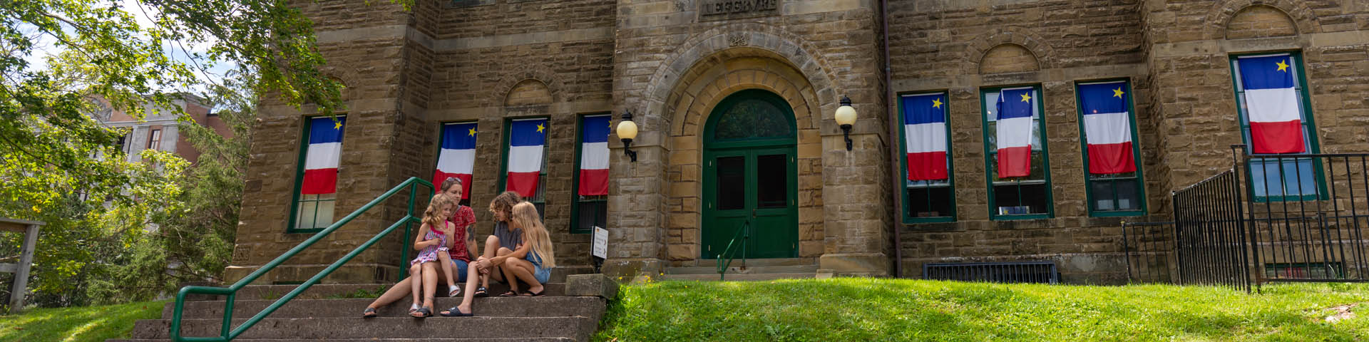A family sits on the front steps of the Monument-Lefebvre building. Acadian flags are hanging on the windows of the building.