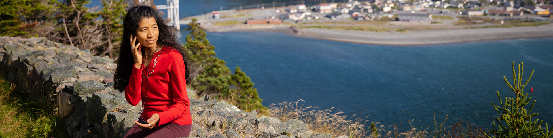 a person listening to an audio device, sits on a stone wall overlooking a coastal town