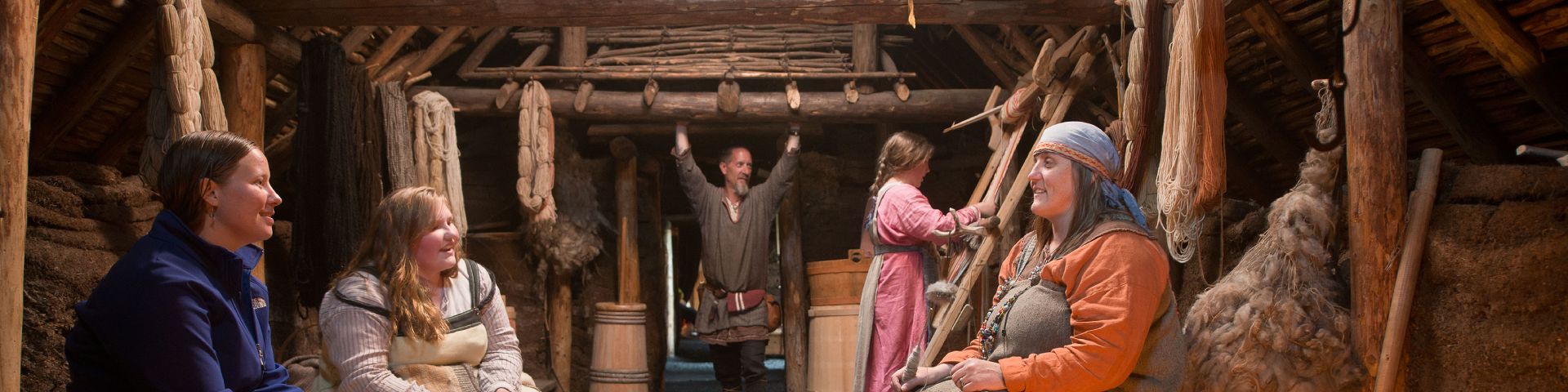 4 Parks Canada interpreters in Viking costume sit in a wooden building with a visitor at L’Anse aux Meadows National Historic Site