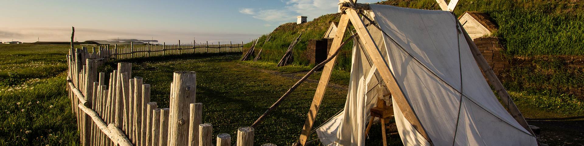 A view of a canvas tent in front of a wood building covered in grass from the Viking encampment at L'Anse aux Meadows National Historic Site