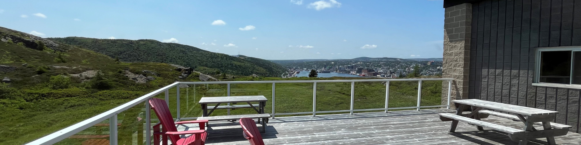 A wooden deck is seen with two red chairs and a wooden picnic table with a view of hills and a cityscape in the background.