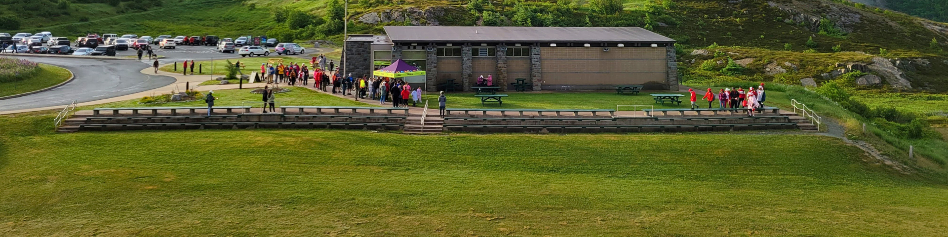 A wide photo shows a big open field with a stone building in the centre behind it and three levels of wooden bench seating along the field's edge.