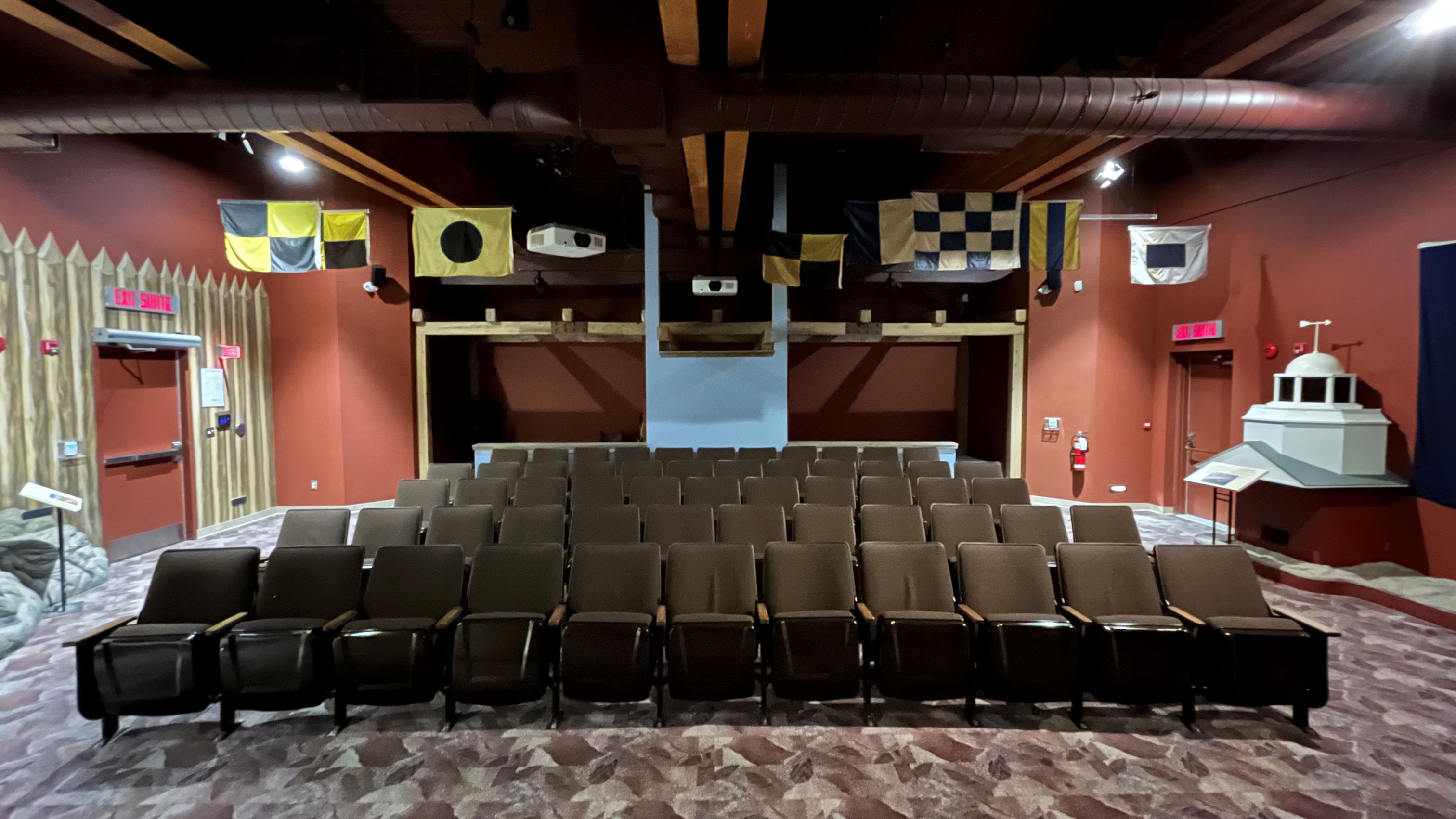 Five rows of theatre seating are seen from the stage perspective. Several flags are hanging from the ceiling, and there is a door on either side of the room.