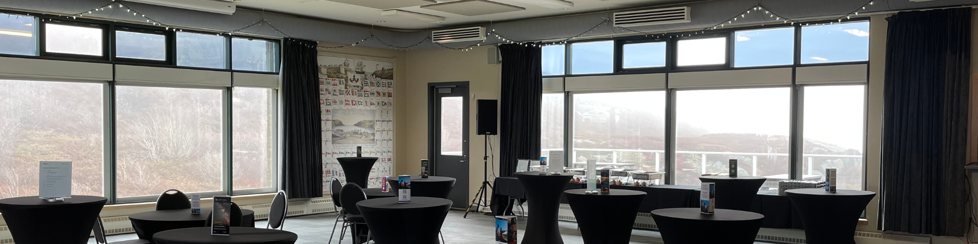 A wide photo that shows an open space in a room with three windows that have curtains opened and throughout the room are several tall small tables setup.